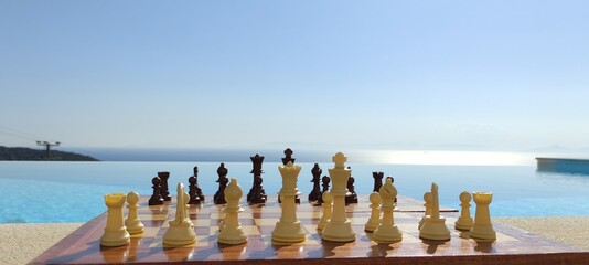 A chessboard, in front of an infinity pool, overlooking the sea, during a sunny, summer day.
