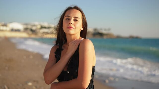 Portrait of beautiful confident Caucasian woman in black dress posing in sunlight on sandy beach. Front view of slim young lady looking at camera with turquoise Mediterranean sea waves at background