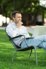 Freelancer man with laptop working in garden sit in chair on grass outdoors. Young blogger male work on computer in public park processes video for social media content or talking in video call.