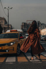 A brunette woman in an autumn long leather coat crosses the road in the city with yellow taxi....