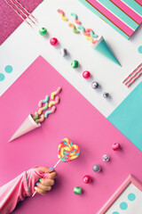 Collection of party objects and sweets on geometric paper flat lay. Chocolate, candy and rainbow lollipop swirl in hand. Geometric layered background in bold pink and mint blue on white background.