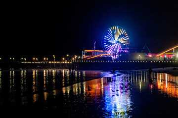 The lights of the Santa Monica Pier, the Ferris wheel and the Rollercoaster reflected in the water of the Pacific Ocean on Santa Monica Beach.