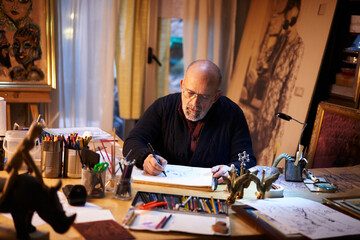 Artist working in the studio. Older man painting a picture using a magnifying glass in a creative and chaotic environment.