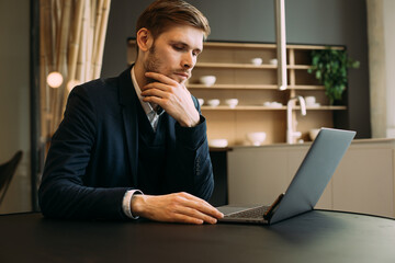 Confident businessman working remotely from a home kitchen with a laptop and notebook. Side view of a young man in smart casual wear making notes during zoom video meeting conference call remote