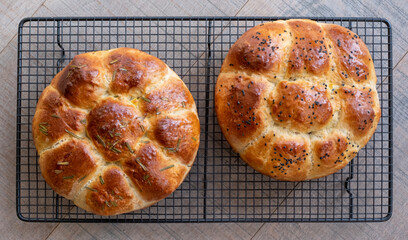 Two round share and tear traditional home cooked Jewish Challah bread loaves, on a wire tray. One is topped with Nigella seeds and the other with rosemary. 
