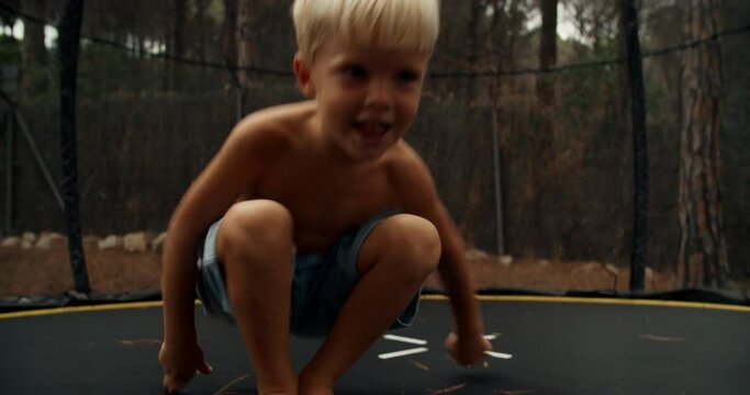 Happy smiling caucasian kid jumps on trampoline on the fresh air. Child makes a U-turn jump on a trampoline in the forest. Cute little boy enjoys his free time and frolics on a warm summer day.