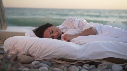 Happy young smiling woman sleeping in white soft bed with turquoise sea waves splashing with foam on sandy beach at background. Portrait of carefree Caucasian lady with closed eyes lying outdoors