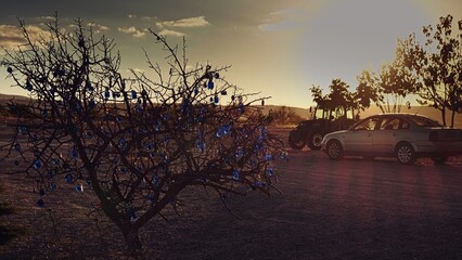 Defocused glowing Evil Eye charms, turkish blue glass beads hanging on wish tree in bright sundown backlight with silhouettes of parked cars on background to enjoy amazing sunset. Cappadocia,Turkey.