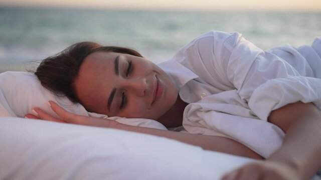 Close-up portrait of happy rested young woman opening eyes waking up smiling with blurred blue sea ocean waves at background. Beautiful Caucasian lady enjoying leisure on beach in sunshine
