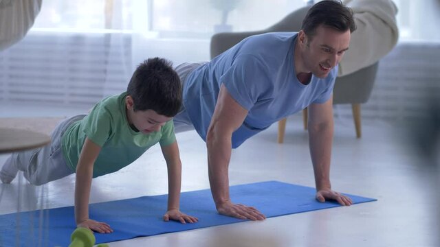 Caring father teaching son to do sport exercises, skinny boy learning to do push ups from floor. Athletic dad doing push-ups with ease while son has hard time, parenting, joint sport activity concept