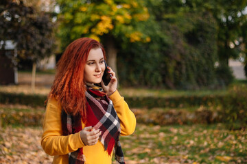 Plakat Young red-haired woman in yellow sweatshirt talking on phone. Lady walking in autumn park on sunny day.