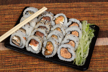 Pieces of sushis, uramaki, with cucumber salad aside and a pair of hashi, on black plastic plate