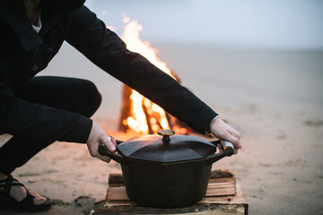 Woman kneels down to lift up a dutch oven while cooking a meal over a fire on the coast of Oregon.