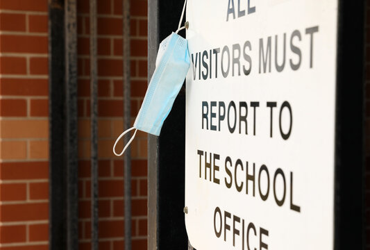 An Authentic Sign At The Entrance To A School Ground A Sign Stating Visitors Must Report To The Front Office With A Blue Protective Face Mask Hanging Off It.