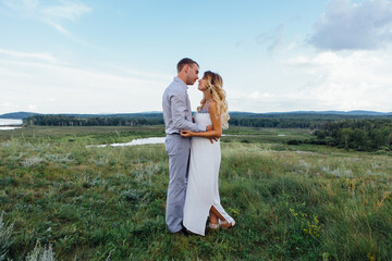 Happy young couple, expecting a baby, walking in summer rainy day on a field. Man hugs his pretty pregnant woman in a white dress. Waiting for the baby, pregnancy concept.