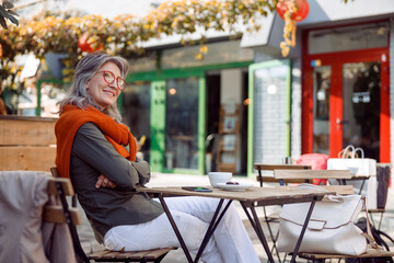 Thoughtful senior lady with crossed arms sits at table with coffee and mobile phone on outdoors cafe terrace on autumn day