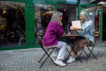 Fototapeta na wymiar Excited senior Asian woman opens gift presented by best friend sitting at small table on outdoors cafe terrace on autumn day