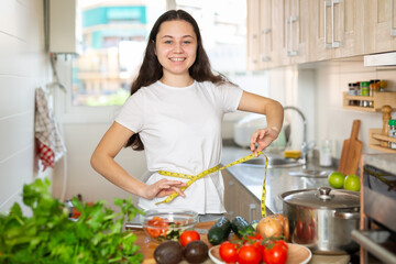 Young pretty girl measuring her waist with measuring tape in the kitchen. Diet and weigh loss concept