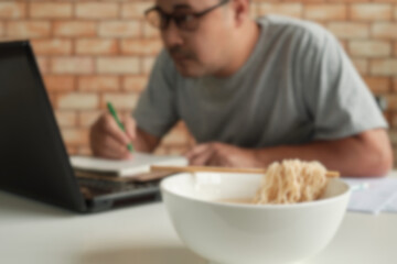 Fototapeta na wymiar Blur focus shot, Thai male worker busy working with laptop, use chopsticks to eat instant noodles during office lunch breaks, because quick and tasty. Over time Asian fast food, unhealthy lifestyle.