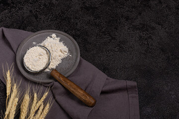 A strainer with a wooden handle with flour in a concrete plate on a black background. Ears of wheat and a linen napkin