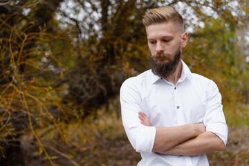 Naklejka na ściany i meble Portrait of pensive bearded attractive young man in white shirt looking over an autumn park in october. Fashionable confident man enjoying a walk in autumn nature during the daytime.