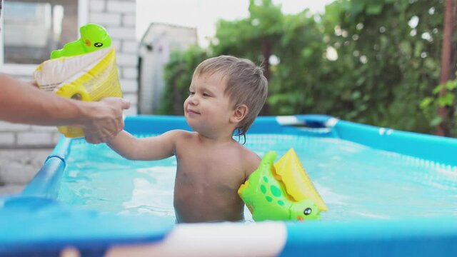 Mom puts a sleeve on the arm of a naked baby who plays in the pool in the yard