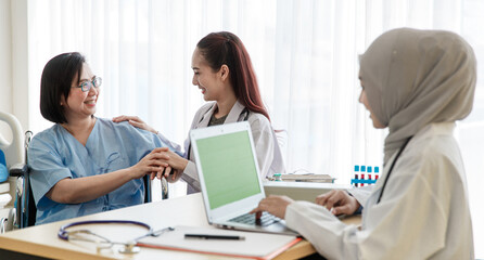 Asian female doctor monitoring checking up senior woman patient on wheelchair while Muslim Arab Islam wears hijab in white lab coat uniform with stethoscope typing information in laptop computer