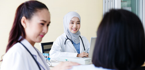 Fototapeta premium Close up shot of Muslim Arab Islam wears hijab in white lab coat uniform with stethoscope typing information in laptop computer while Asian female doctor talking with patient in blurred foreground