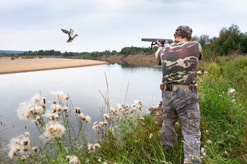a hunter stands on the river bank and shoots a flying duck