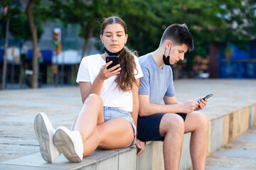 Two teenagers in masks are sitting and watching in their smartphones