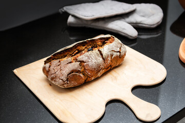 Rustic baked sourdough bread on cuttings board placed on black kitchen counter. Oval fresh yeast bread loaf made with rye and wheat flour and nuts From scratch made sourdough bread. Selective focus.