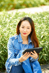 Pretty young Vietnamese student in denim clothes sitting on campus with digital tablet in hands, reading article for upcoming class