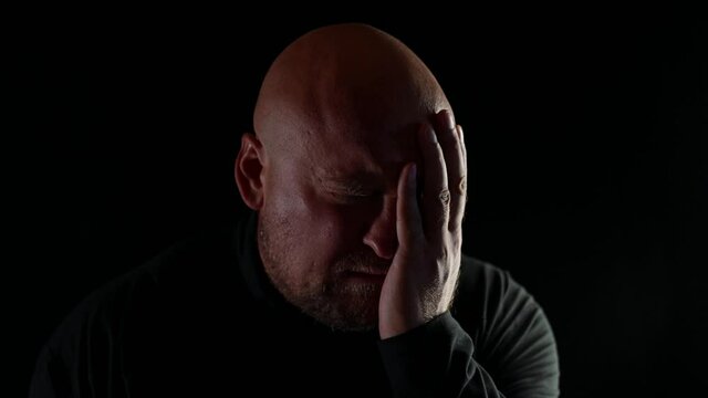 Portrait of a bald man in pain touching his face with his hand on a black background in the dark