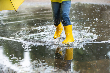 A little girl with a yellow umbrella and rubber boots jumps merrily through the puddles, splashes are flying in the sunbeams.