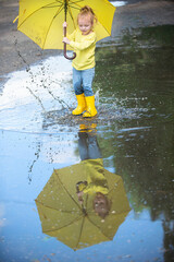 A little girl with a yellow umbrella and rubber boots jumps merrily through the puddles, splashes are flying in the sunbeams.