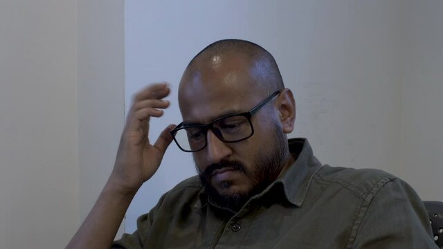 Ethnic Minority Bald Male Wearing Glasses Tapping Forehead With Fingers In Chair. Static Shot
