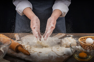 Women's hands, flour and dough. A woman in an apron cooking dough for homemade baking, a rustic home cozy atmosphere, a dark background with unusual lighting.