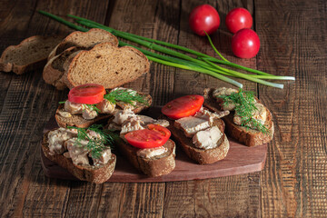 On a wooden background, there are ready-made sandwiches with cod liver, tomato and greens, canned fish are lined in a dark plate. 