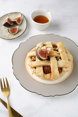 A sweet fig tart pastry on a sophisticated plate, two golden dessert spoons, honey, cut figs on a marble surface