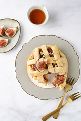 A sweet fig tart pastry on a sophisticated plate, two golden dessert spoons, honey, cut figs on a marble surface, top view