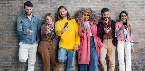 Interracial group of young people joking together using smartphones and browsing social network content online standing against a brick wall. Technology concept.