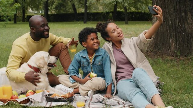 Beautiful Afro-American family smiling and posing for smartphone camera while taking selfie with dog on picnic in park