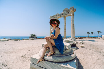 A beautiful girl in a dress, hat and sunglasses poses with a view of the ruins of the ancient Roman temple of Apolon. Side, Turkey.