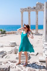 A beautiful girl in a dress, hat and sunglasses poses with a view of the ruins of the ancient Roman temple of Apolon. Side, Turkey. Vertical photo.