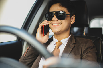 Smiling Asian businessman in sunglasses is driving a car while doing conversation on smart phone