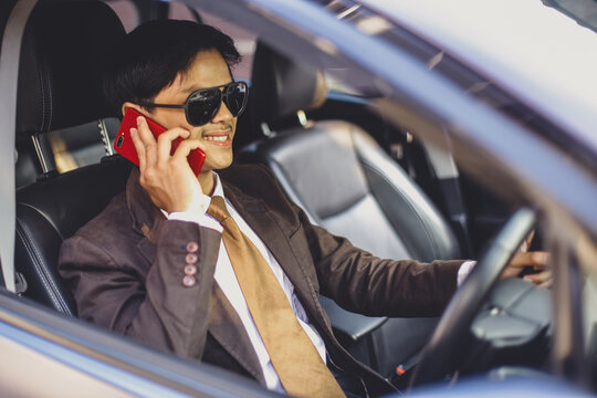 Smiling Asian businessman in sunglasses is driving a car while doing conversation on smart phone