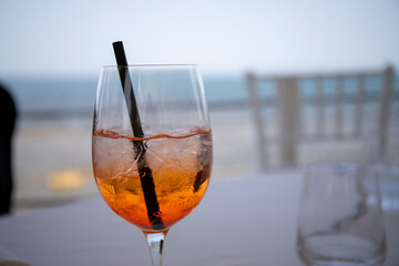 A glass of Spritz (orange alcoholic drink) in the foreground on the white table and the sea in the background.
