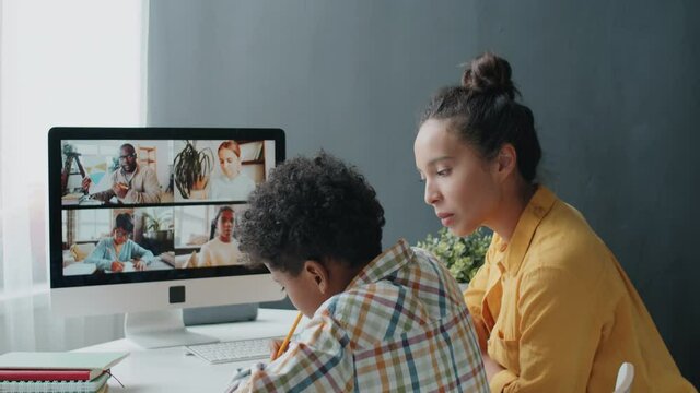 Little Afro-American Boy Watching Online School Lesson Via Video Call On Computer And Taking Notes While Studying At Home With Help Of Mother During Covid-19 Lockdown