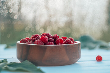 Partially blurred wooden round bowl full of ripe red juicy raspberries on bokeh background with raspberry bush leaves