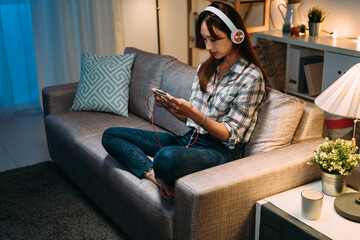Obraz premium asian girl sitting on couch in casual wear is holding and watching online videos on her smartphone with headphone during nighttime in a cozy living room at home